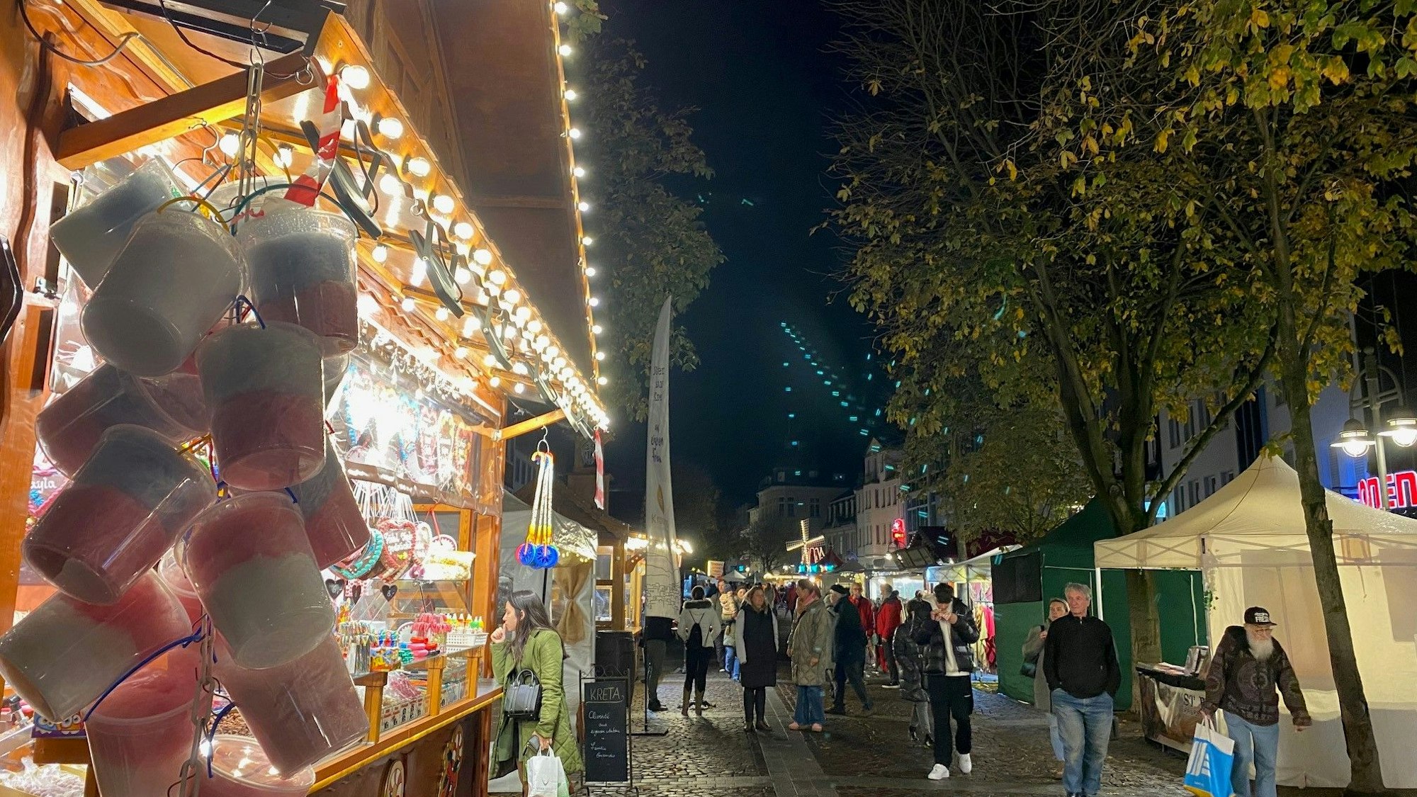 Weihnachtsmarktbuden aus Holz mit geschmückten Dächern reichen sich über den Marktplatz. Menschen schlendern zwischen ihnen umher.
