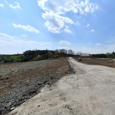 Ein leeres Feld aus Sand und Erde, in der Mitte ein Weg und ein Mülleimer. Im Hintergrund einige laublose Bäume vor strahlend blauem Himmel.