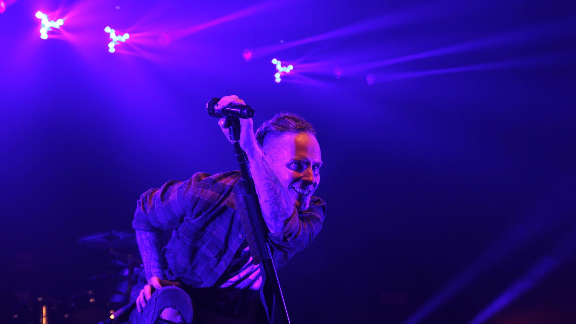 Sänger Corey Taylor kniet mit diabolischem Grinsen neben seinem Mikrofonständer. Die dunkle Beleuchtung verstärkt den Effekt.