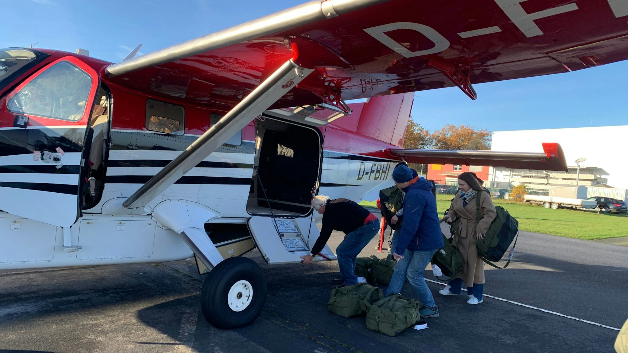 Drei Menschen stehen vor einem kleinen Flugzeug und beladen es mit gepackten Rucksäcken aus olivgrünem Stoff.