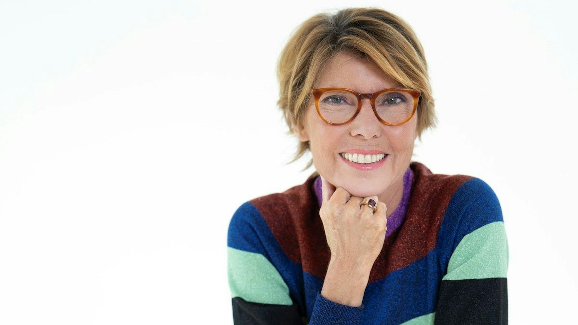 Das Bild zeigt Bettina Böttinger. Die Moderatorin stützt ihr Kinn mit der Hand. Sie trägt eine Brille und einen bunt-gestreiften Pullover.