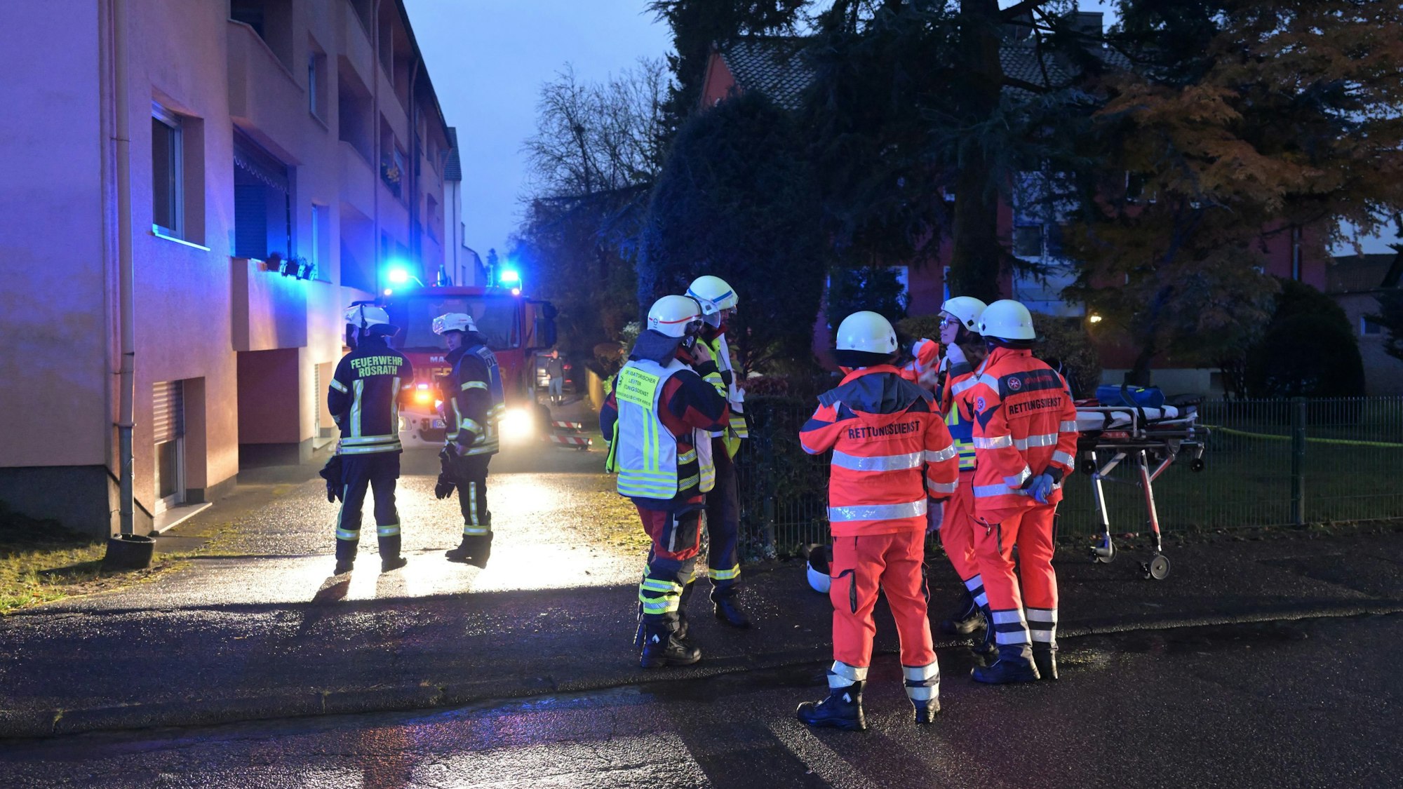 Feuerwehrleute stehen am Abend mit Blaulicht an ihren Einsatzfahrzeugen vor einem Mehrfamilienhaus.