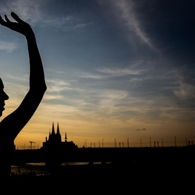 Ein Schatten einer Tänzerin vor blau-goldenen Himmel mit Sonnenuntergang, im Hintergrund die Silhouette des Doms.