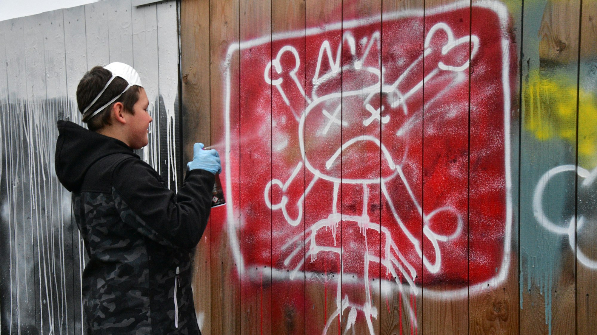 Junge sprüht an weißen Totenkopf auf rotem Grund an einen Holzwand.