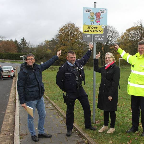 Ordnungsamtsleiter Ferdi Hoss, Schulleiterin Kirsten Schäfer sowie Thomas Claßen und Bernd Schmitz vom Bezirksdienst der Polizei zeigen auf ein Schild mit zwei Schulkindern und der Aufschrift Elternhaltestelle.