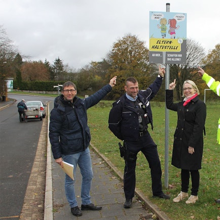 Ordnungsamtsleiter Ferdi Hoss, Schulleiterin Kirsten Schäfer sowie Thomas Claßen und Bernd Schmitz vom Bezirksdienst der Polizei zeigen auf ein Schild mit zwei Schulkindern und der Aufschrift Elternhaltestelle.