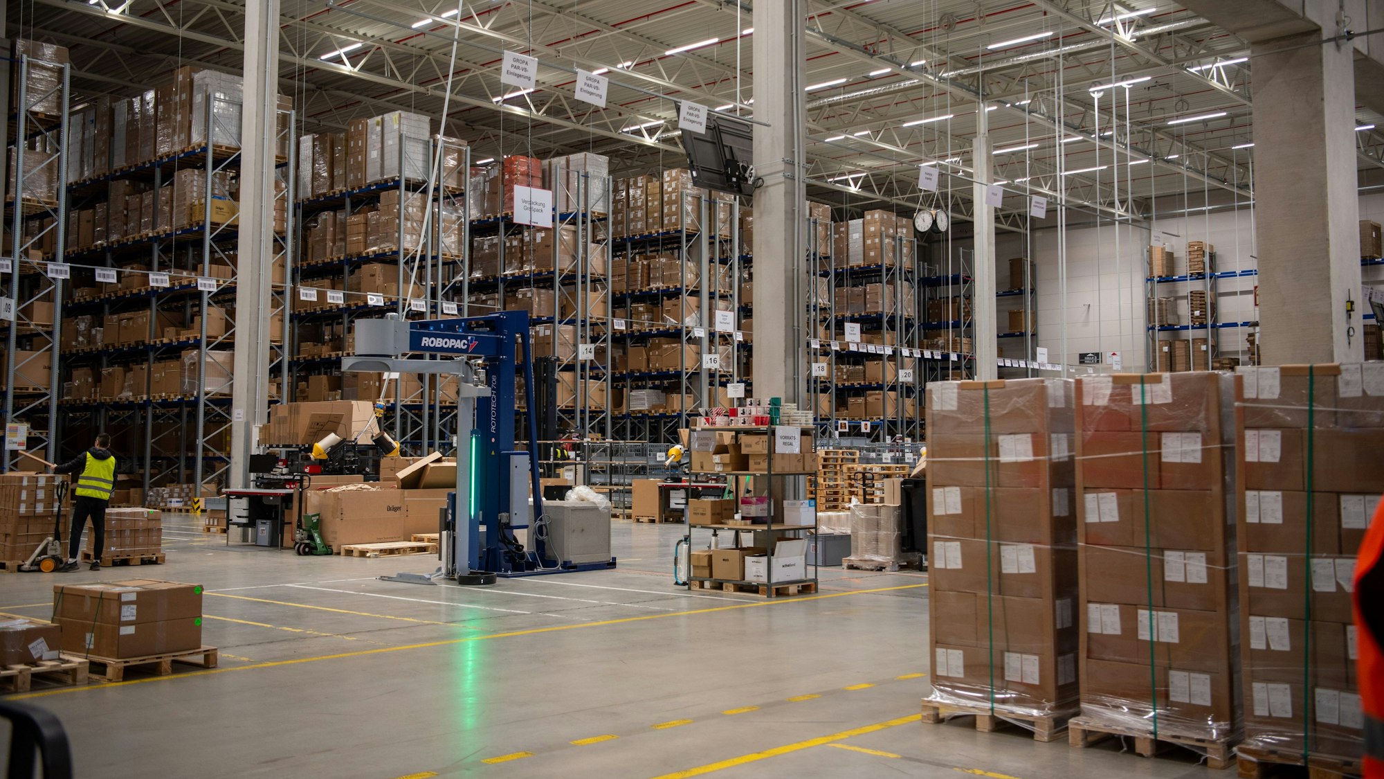 Kartons lagern in großen Regalen und auf Paletten in einer hohen Halle.