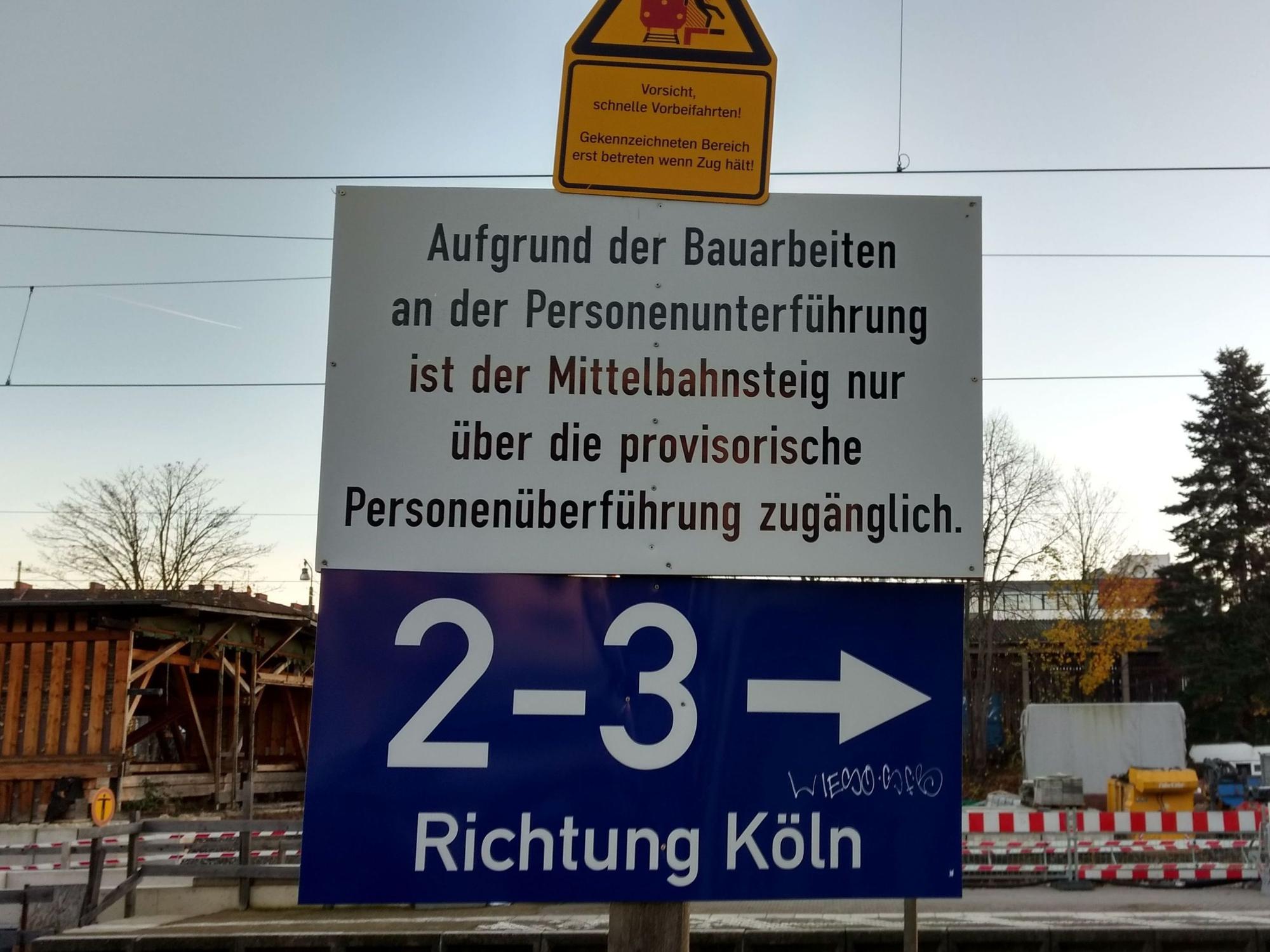 Ein Schild mit der Aufschrift: „Aufgrund der Bauarbeiten an der Personenunterführung ist der Mittelbahnsteig nur über die provisorische Personenüberführung zugänglich.“ Im Hintergrund die Absperrung einer Baustelle.