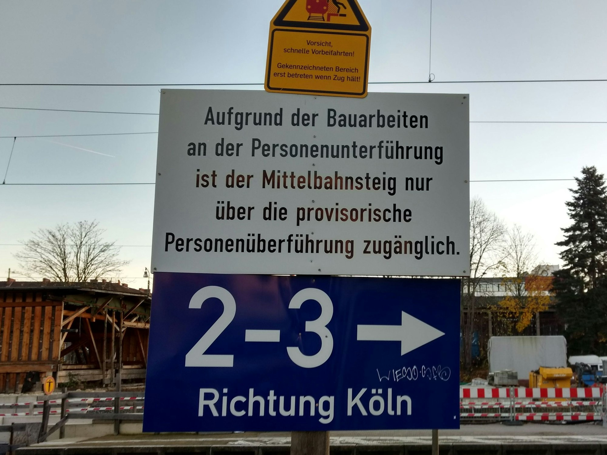 Ein Schild mit der Aufschrift: „Aufgrund der Bauarbeiten an der Personenunterführung ist der Mittelbahnsteig nur über die provisorische Personenüberführung zugänglich.“ Im Hintergrund die Absperrung einer Baustelle.