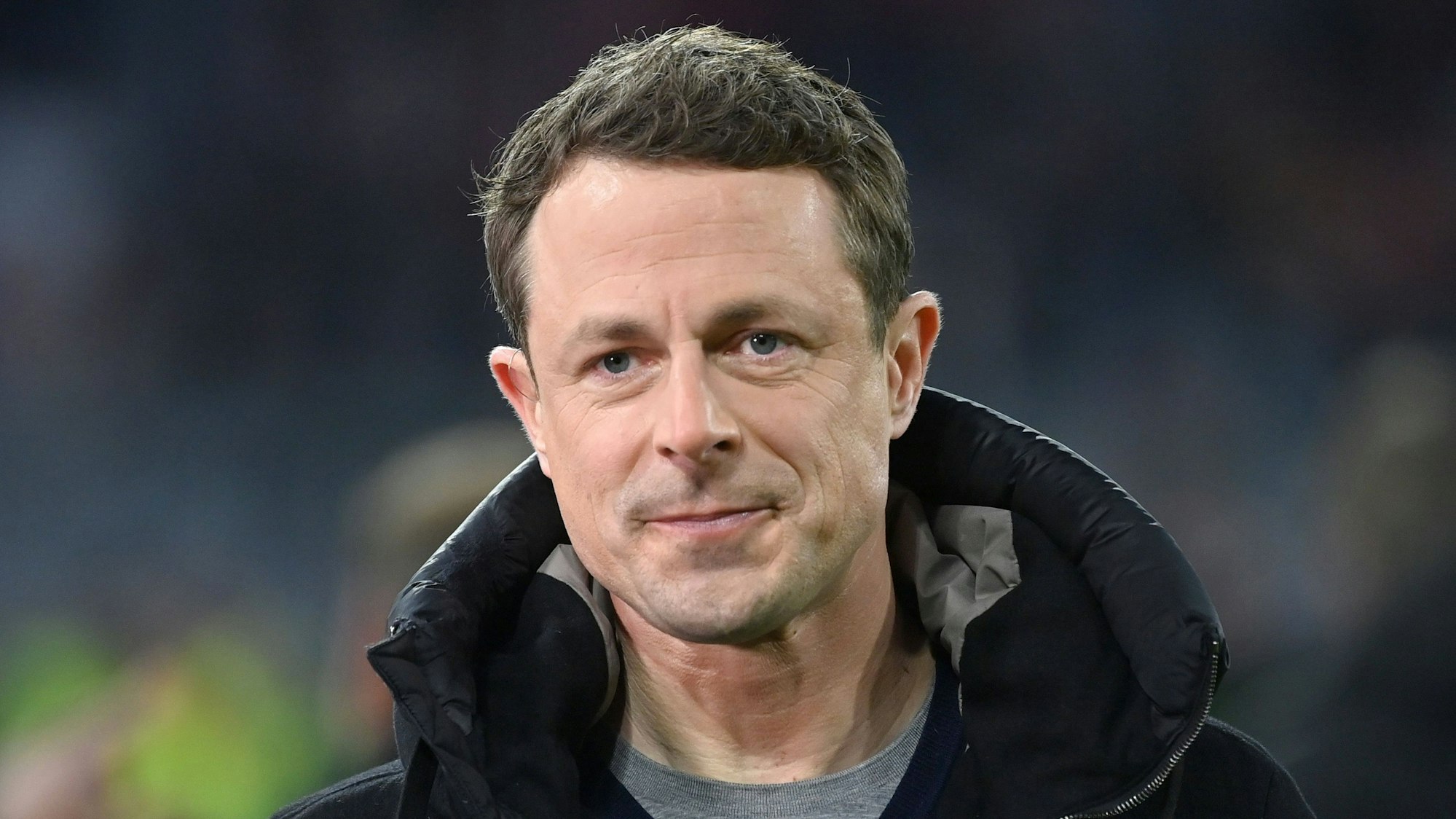 Moderator Alexander Bommes blickt bei einer ARD-Übertragung während eines Fußballspiels in die Kamera. Er trägt eine schwarze Jacke und einen grauen Pullover.