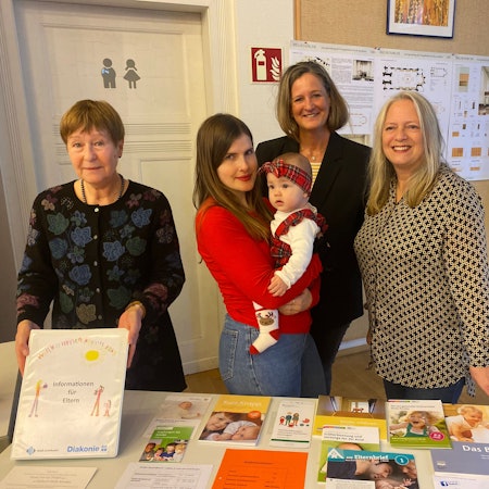 Vier Frauen stehen vor einem Tisch. Eine trägt ein Säugling auf dem Arm.