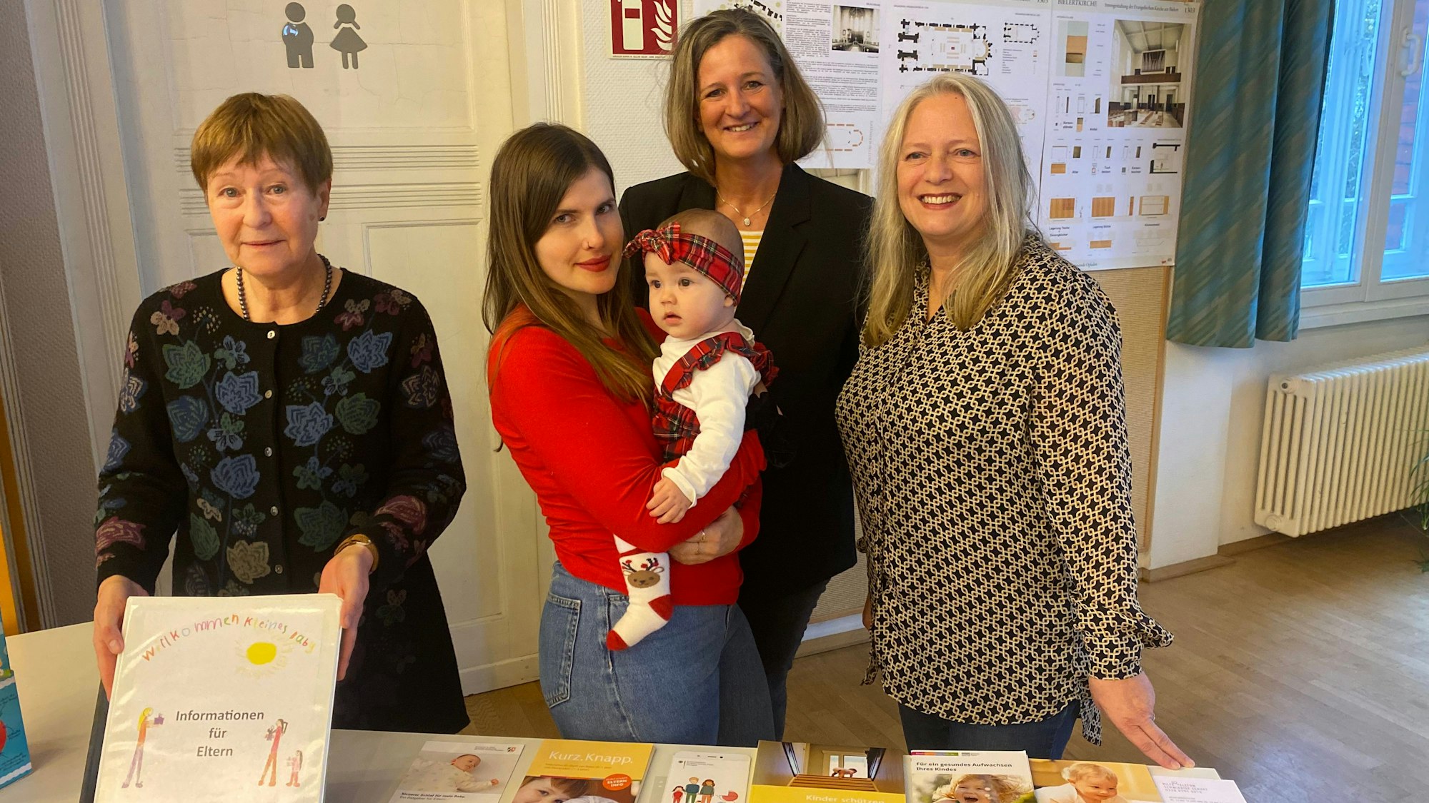 Vier Frauen stehen vor einem Tisch. Eine trägt ein Säugling auf dem Arm.