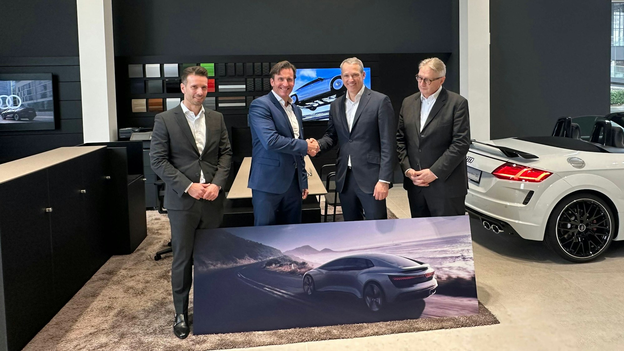 Mehrere Personen stehen nebeneinander, zwei davon geben sich die Hand. Vor ihnen liegt das Bild eines Autos auf der Straße. Im Hintergrund steht rechts ein echtes Auto ohne Verdeck.