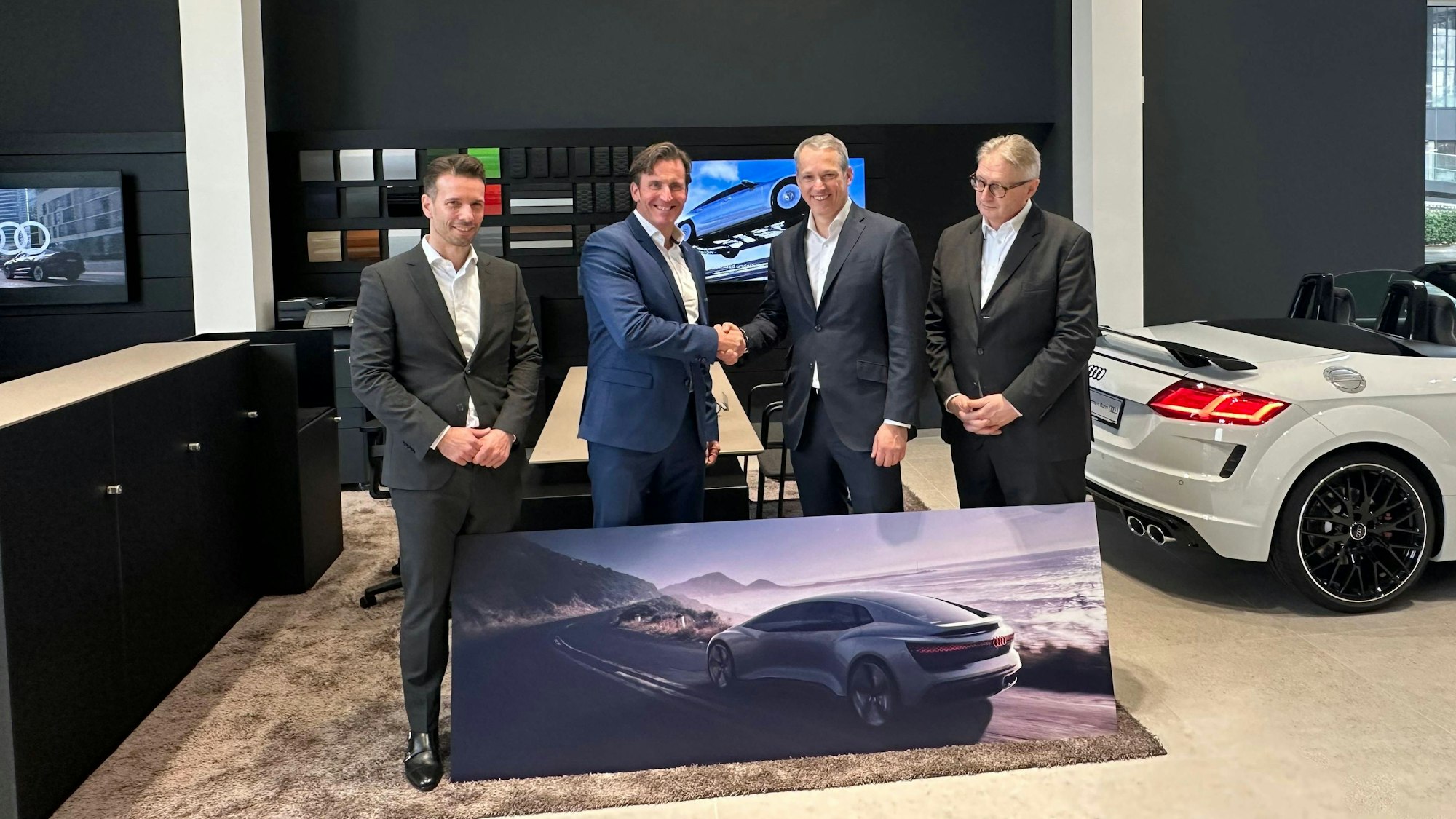 Mehrere Personen stehen nebeneinander, zwei davon geben sich die Hand. Vor ihnen liegt das Bild eines Autos auf der Straße. Im Hintergrund steht rechts ein echtes Auto ohne Verdeck.