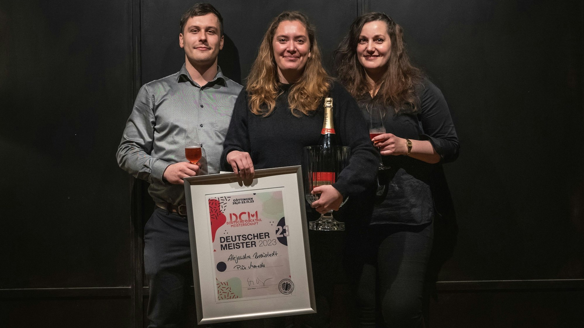 Drei Personen stehen vor einer schwarzen Wand und halten eine Urkunde.