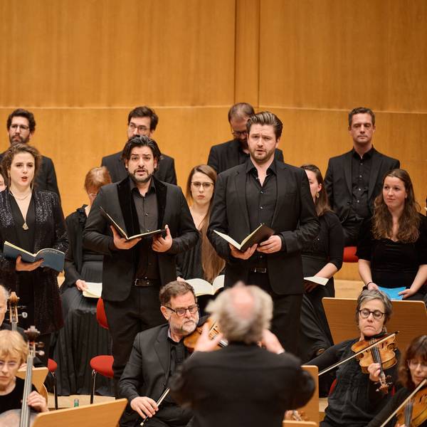 Auf dem Bild sind die vier Solisten zu sehen, vor ihnen das Orchester und hinter ihnen weitere Sängerinnen und Sänger. Alle sind schick angezogen. Im Vordergrund verschwommen der Dirigent Philippe Herreweghe
