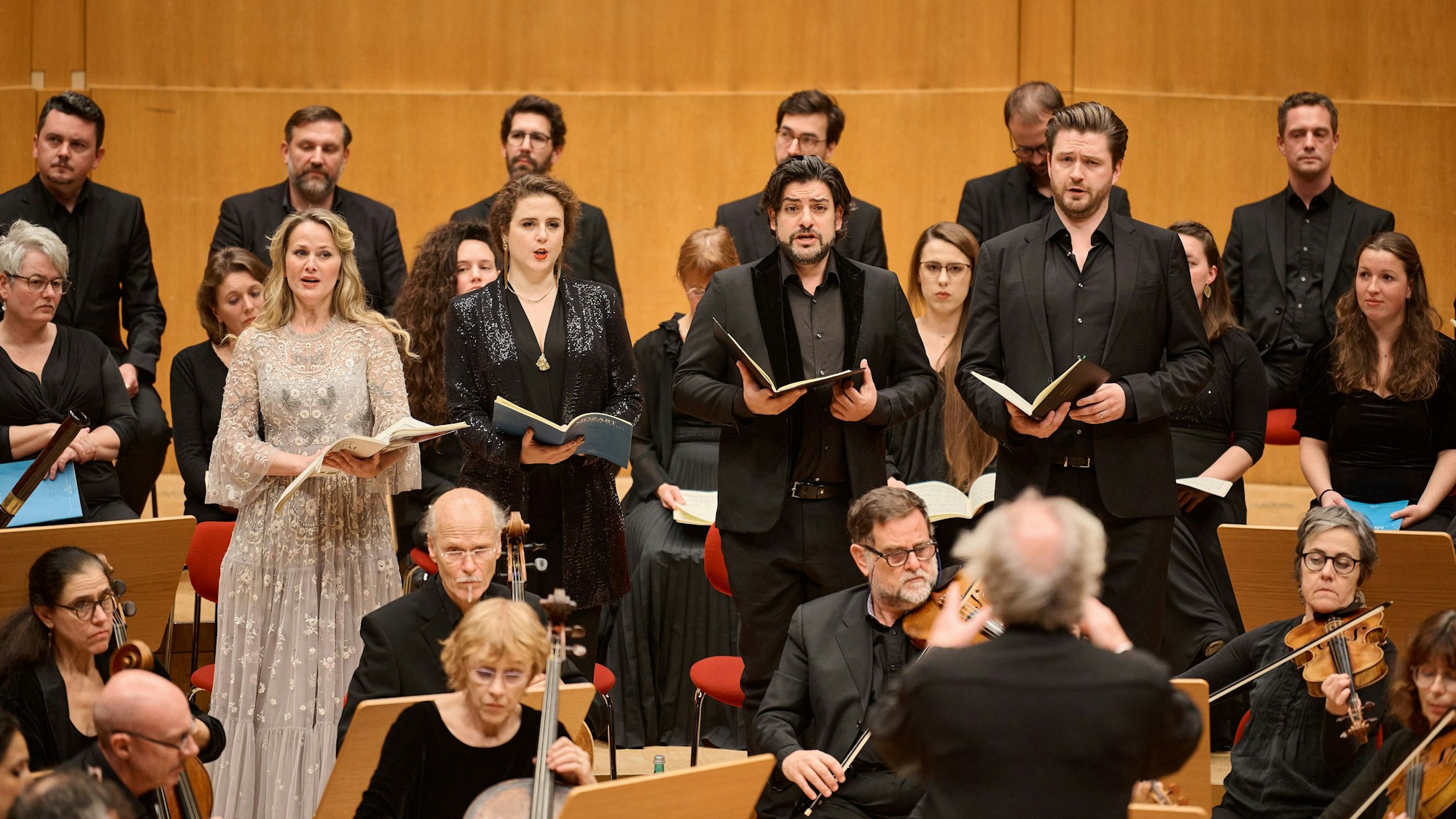 Auf dem Bild sind die vier Solisten zu sehen, vor ihnen das Orchester und hinter ihnen weitere Sängerinnen und Sänger. Alle sind schick angezogen. Im Vordergrund verschwommen der Dirigent Philippe Herreweghe