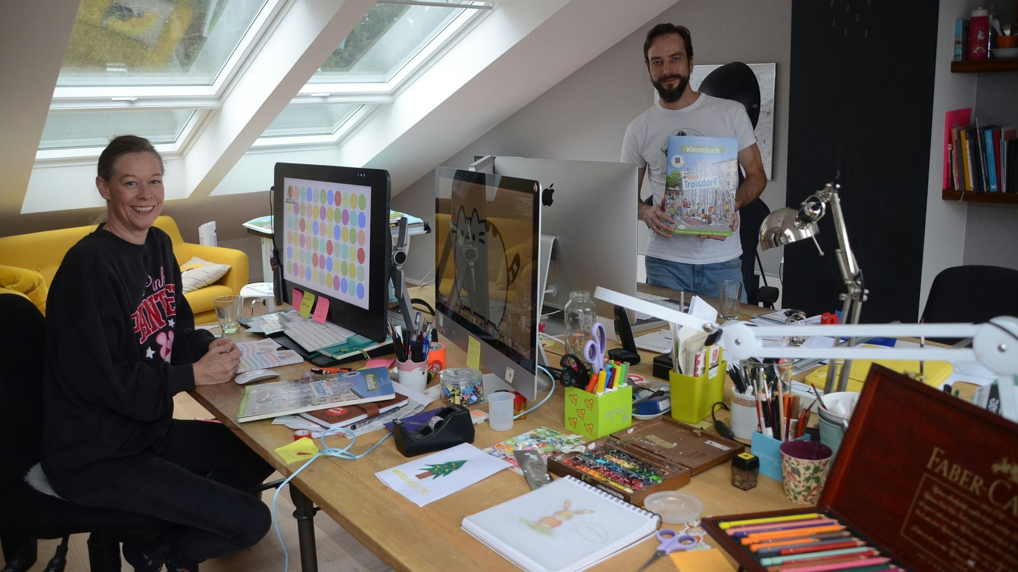 Ein Mann und eine Frau in einem Arbeitszimmer im Dachgeschoss. Durch große Fenster fällt Licht herein; sie trägt ein schwarzes Sweat-Shirt, er ein weißes T-Shirt. Auf den Schreibtischen liegen bunte Stifte und andere Malutensilien.