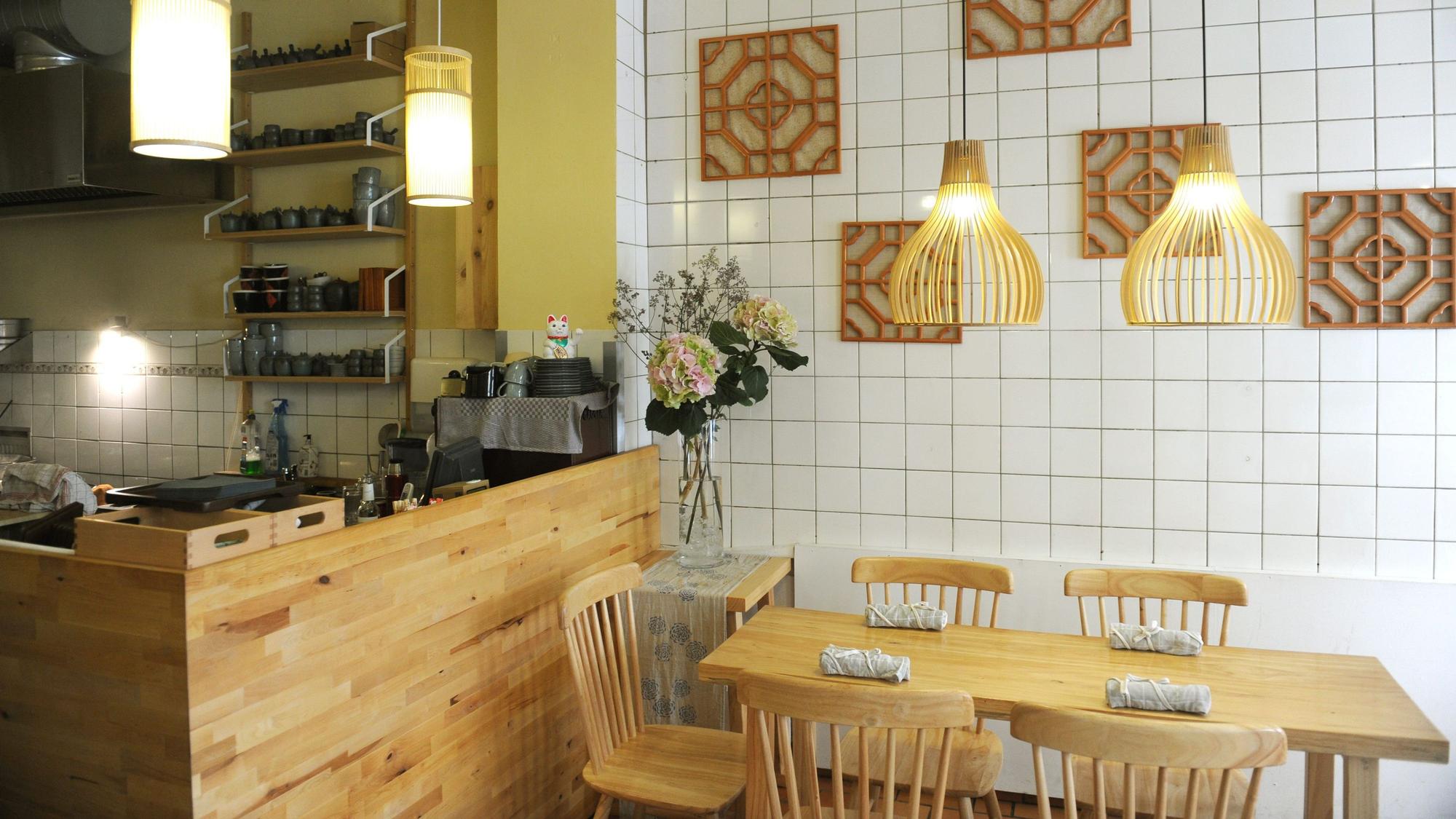 Der Innenraum von Bai Lu Noodles; Holztheke und Holztische vor weißen Kacheln mit modern-asiatischer Holzdeko an der Wand.