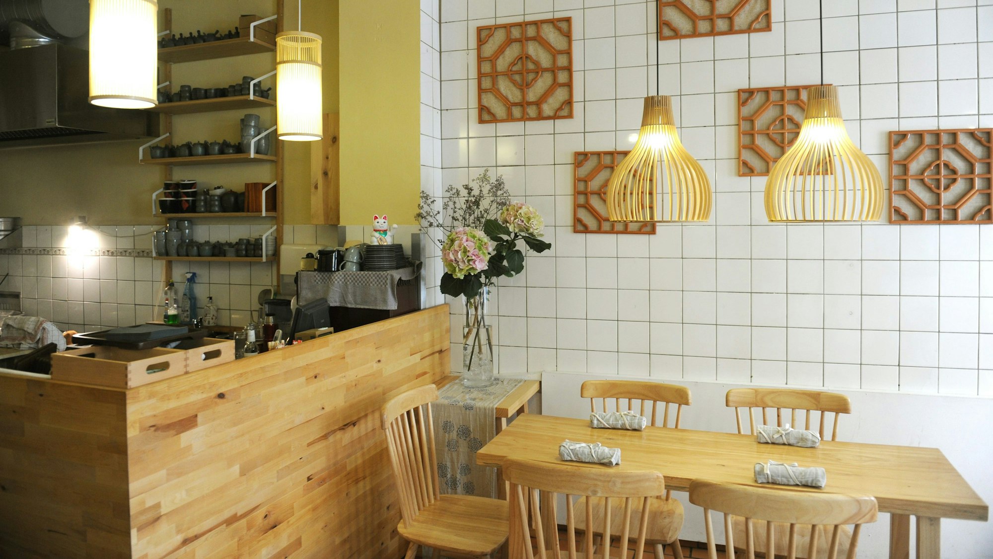 Der Innenraum von Bai Lu Noodles; Holztheke und Holztische vor weißen Kacheln mit modern-asiatischer Holzdeko an der Wand.