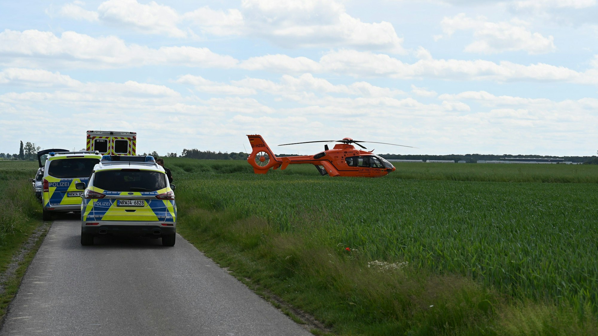 Auf dem Foto sind ein Rettungshubschrauber, der in einem Feld neben dem Vinger Weg gelandet ist, Polizeiwagen und ein Rettungswagen zu sehen.