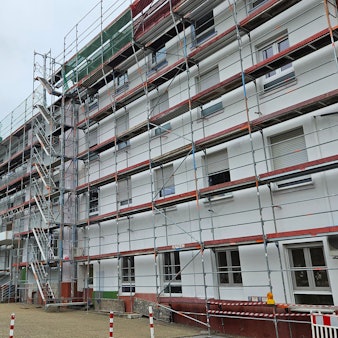Ein großes helles Gebäude mit mehreren Stockwerken ist eingerüstet, im Hintergrund steht eine Backsteinkirche.