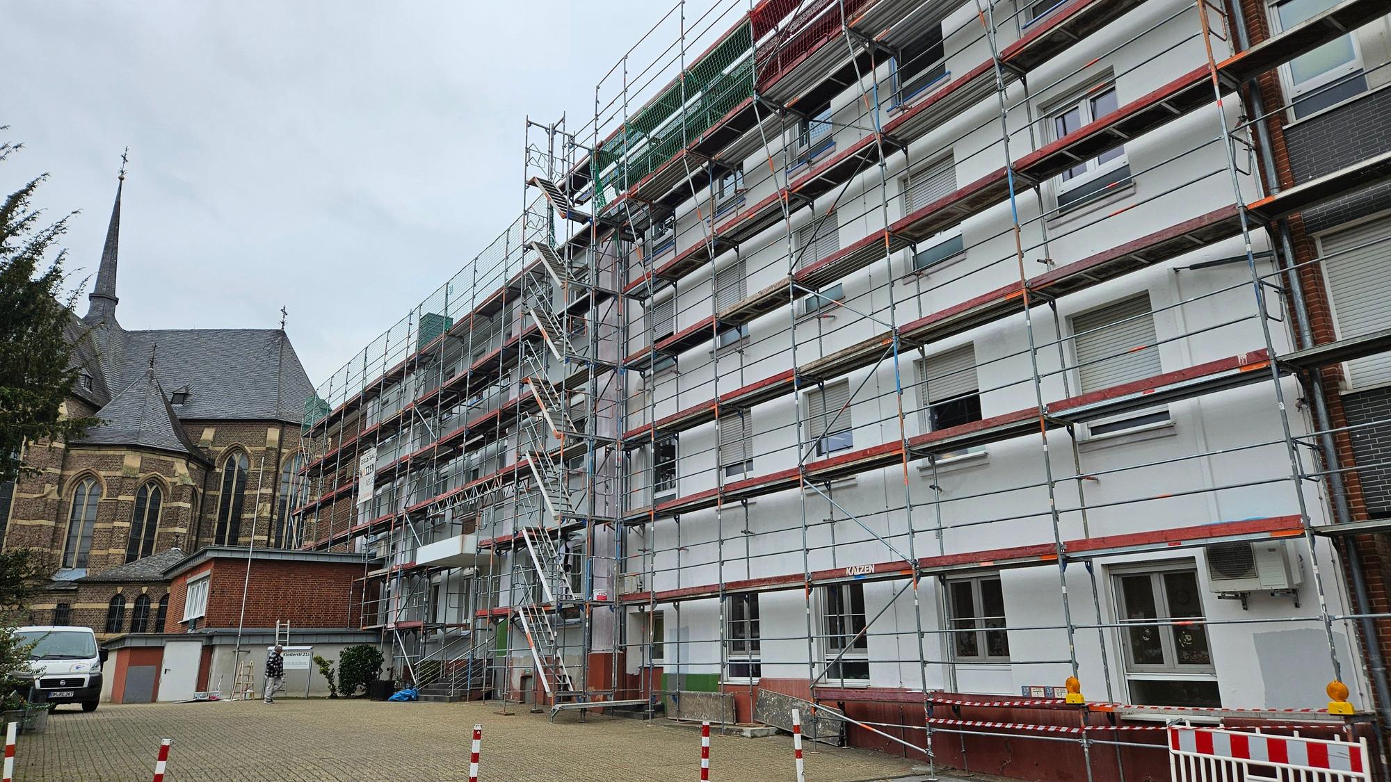 Ein großes helles Gebäude mit mehreren Stockwerken ist eingerüstet, im Hintergrund steht eine Backsteinkirche.