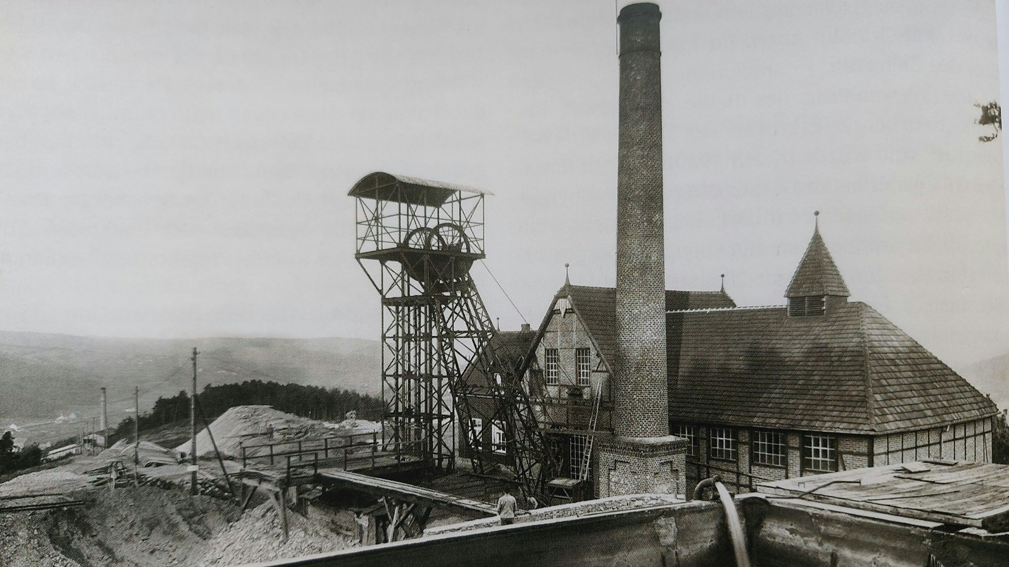 Historische Aufnahme einer alten Grubenanlage mit Förderturm und Schornstein, davor Halden.