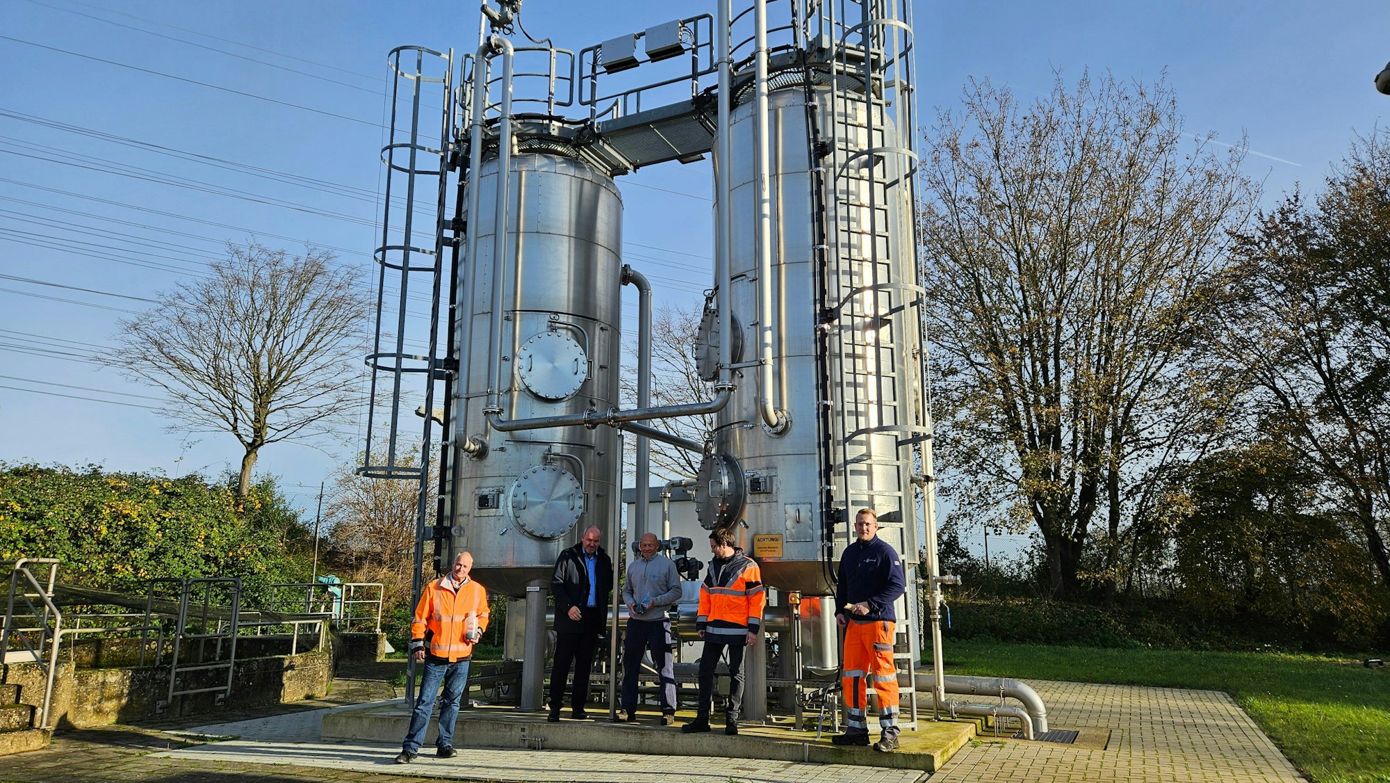 Menschen stehen vor einer Kläranlage mit zwei großen Metall-Türmen.