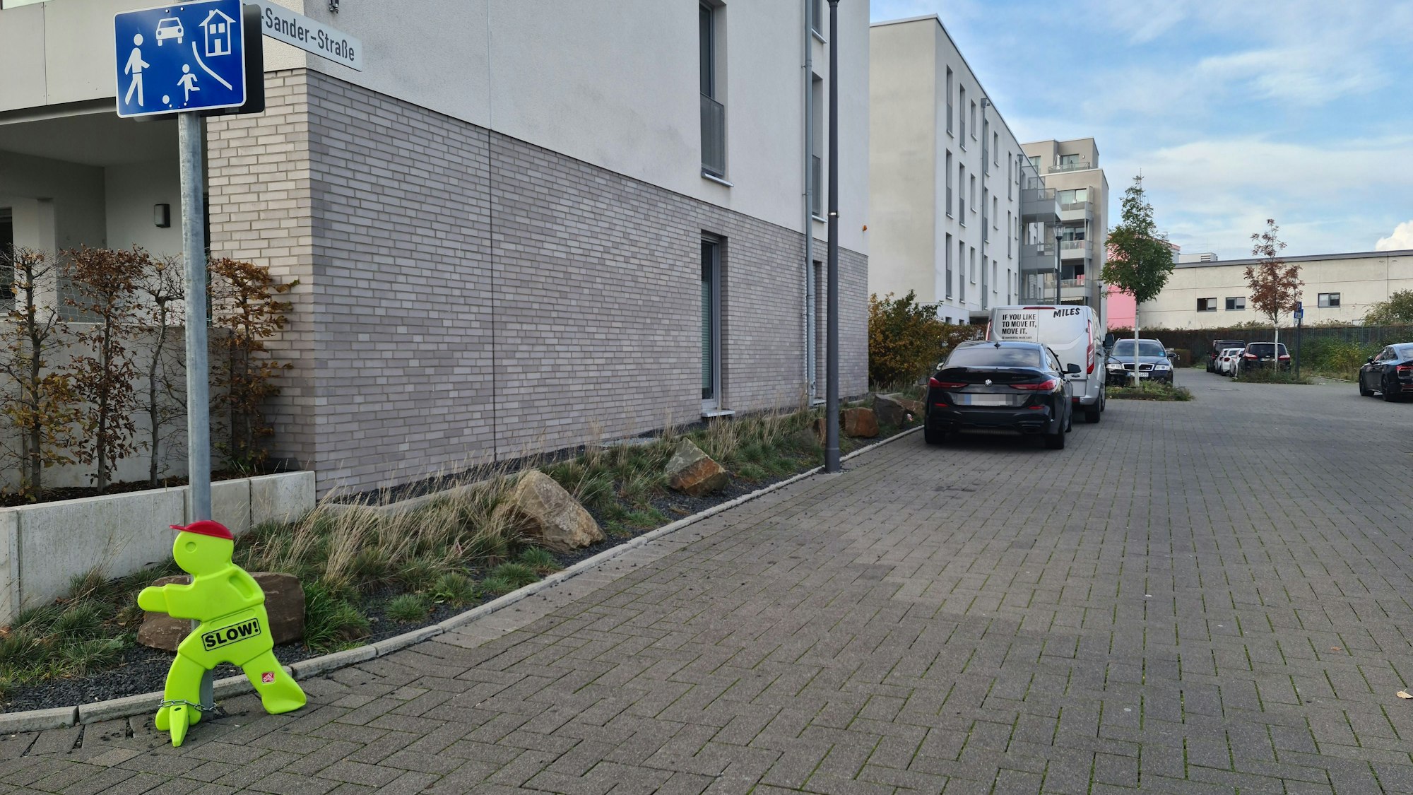 Ein Straßenschild mit dem blauen Zusatzschild, das eine Spielstraße auszeichnet und ein gelbes Plastikmännchen stehen an der Einmündung einer Straße, an der auch einige Autos parken.