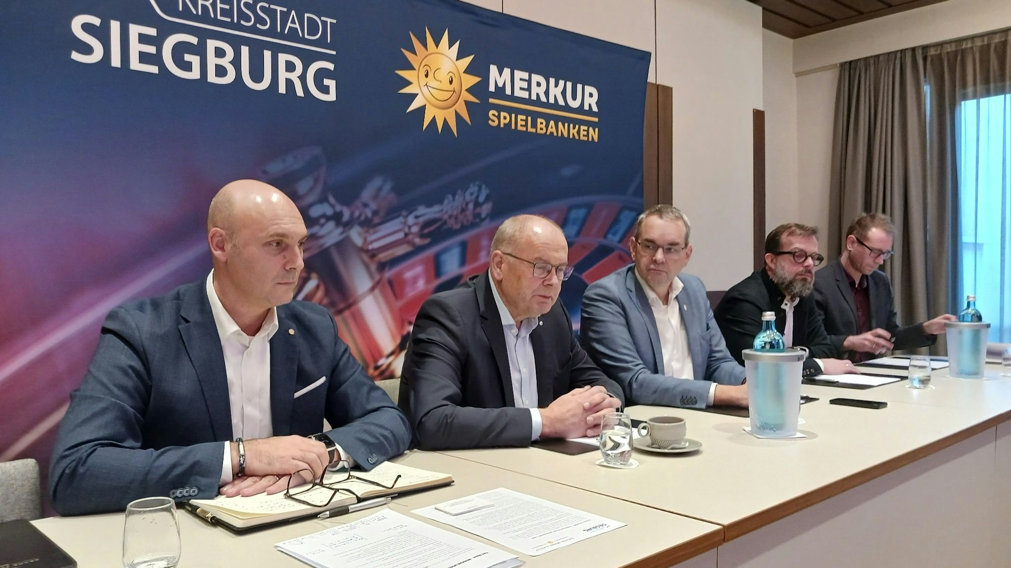 Fünf Mann sitzen an einem langen Tisch vor einem Plakat mit einem Roulette-Tisch