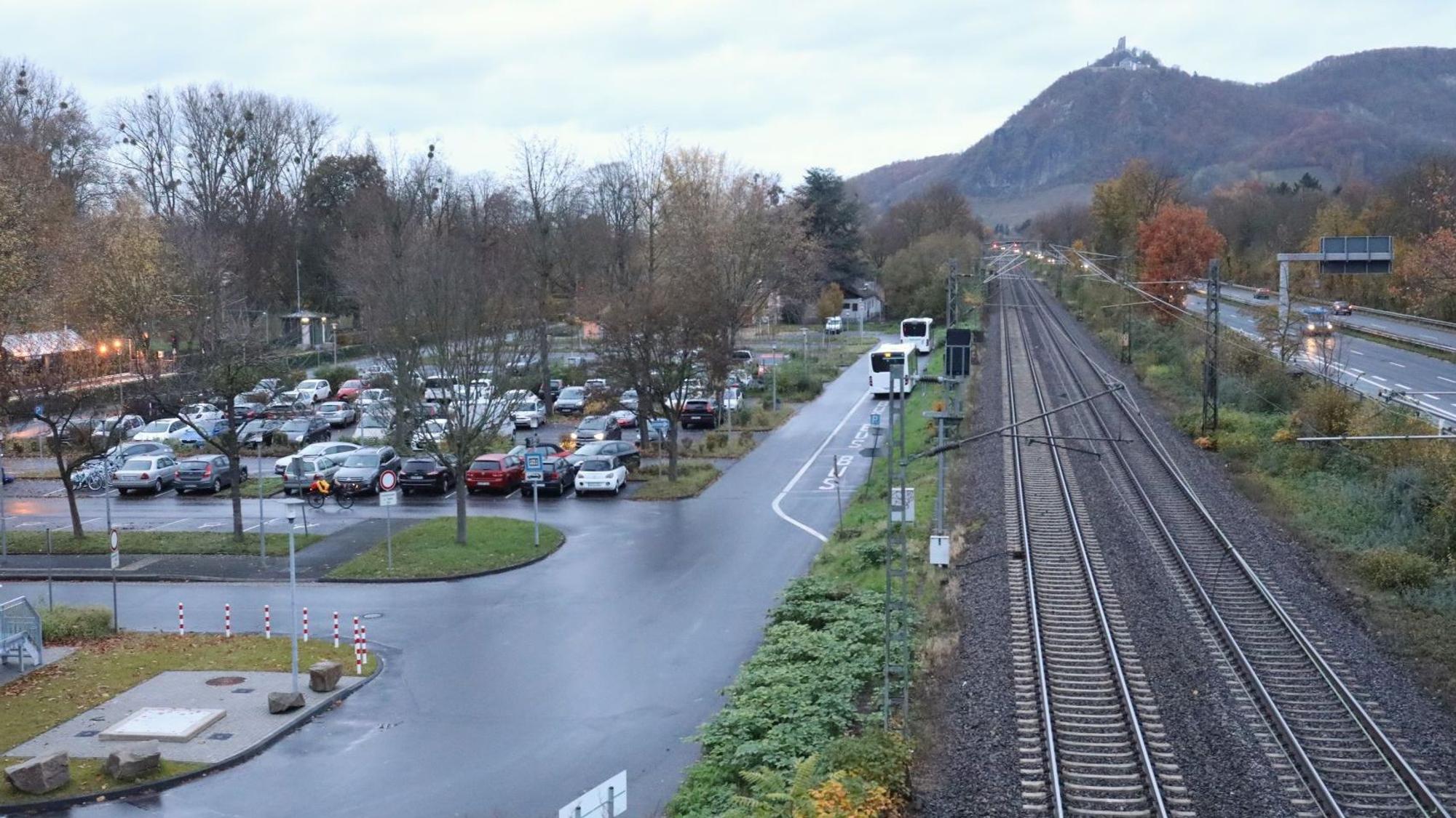 Neben einem Parkplatz verläuft die Bahntrasse, rechts davon befindet sich die Bundesstraße 42. Im Hintergrund ist der Drachenfels zu sehen.