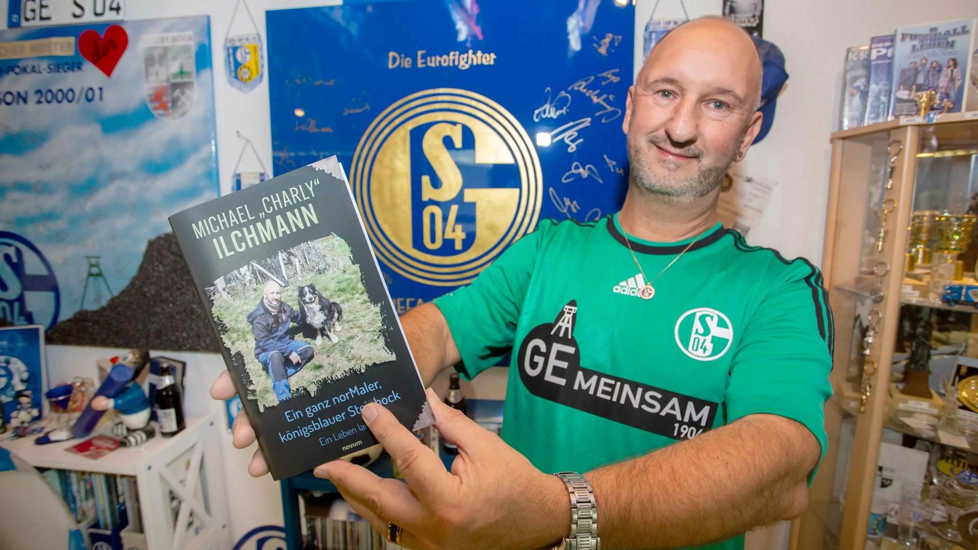 Ein Mann steht in einem Zimmer voller Schalke 04-Fanartikel und hält seine Autobiografie in den Händen.
