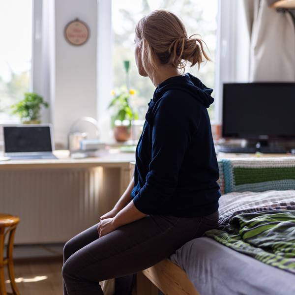 Eine junge Frau sitzt auf dem Bett in ihrem WG-Zimmer und schaut aus dem Fenster (gestellte Szene).