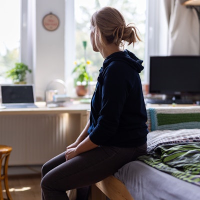 Eine junge Frau sitzt auf dem Bett in ihrem WG-Zimmer und schaut aus dem Fenster (gestellte Szene).