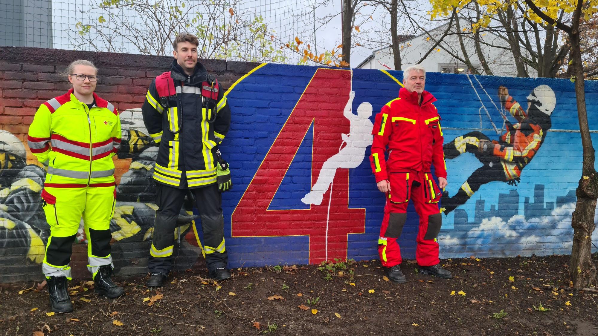 Eine Frau in Notfallsanitäter-Uniform und zwei Männer in Feuerwehr-Kluft stehen vor einer Mauer, die mit Szenen aus dem Alltag der Feuerwehr bemalt wurde.