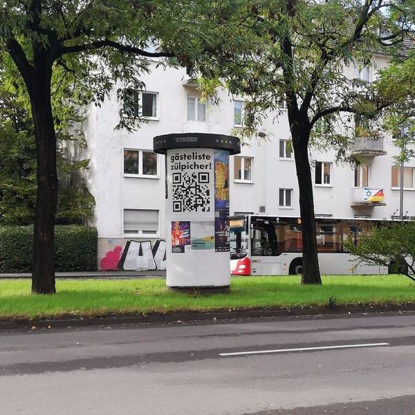 Auf einer Litfaßsäule hängt ein Plakat mit der Aufschrift „Gästeliste Zülpicher“, darunter ist ein großer QR-Code gedruckt.