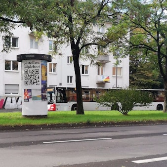 Auf einer Litfaßsäule hängt ein Plakat mit der Aufschrift „Gästeliste Zülpicher“, darunter ist ein großer QR-Code gedruckt.