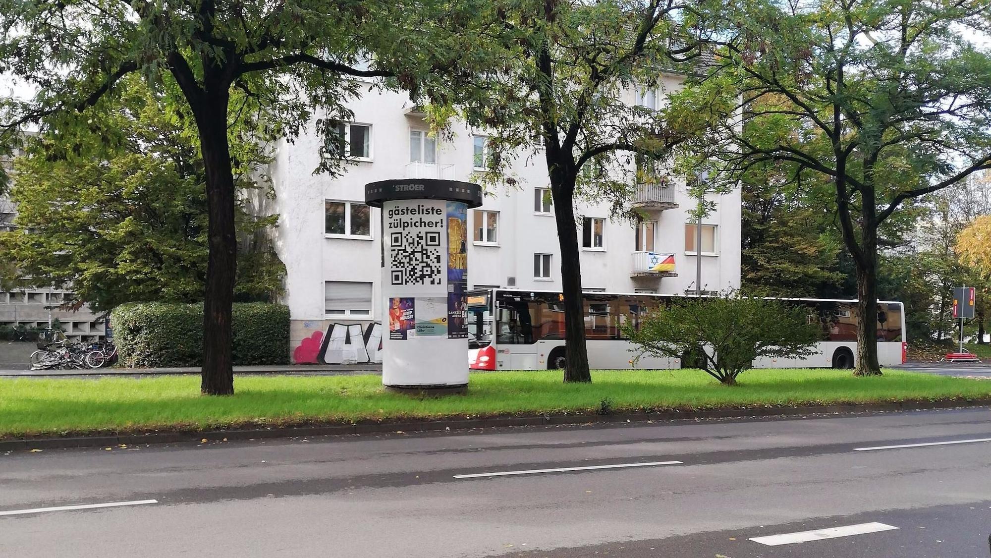Auf einer Litfaßsäule hängt ein Plakat mit der Aufschrift „Gästeliste Zülpicher“, darunter ist ein großer QR-Code gedruckt.