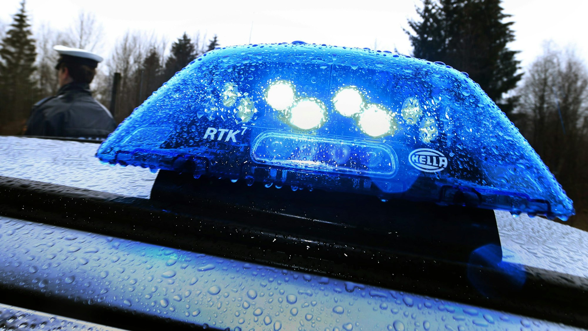 Ein Polizist steht im Regen vor einem Streifenwagen, dessen Blaulicht aktiviert ist. (Symbolbild)