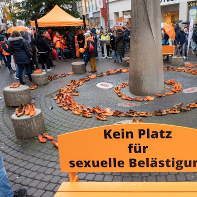 Orange Bank mit der Aufschrift „Kein Platz für sexuelle Belästigung“, im Hintergrund sind orange Schuhpaare kreisförmig aufgestellt