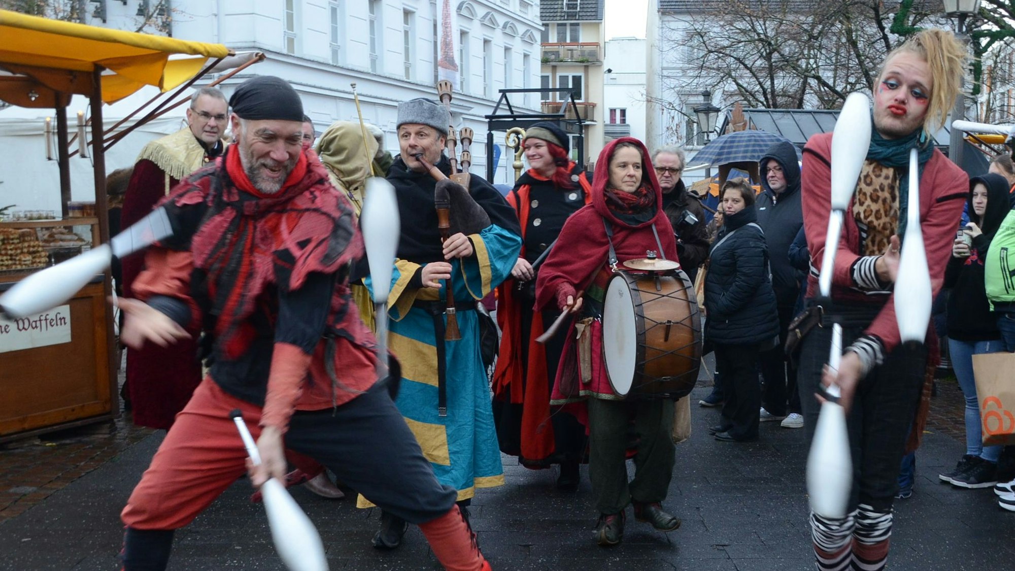 Menschen in mittelalterlichen Kostümen; vorne Jongleure, dahinter ein Mann mit Dudelsacke, eine Frau mit Pauke. Am Rand stehen Zuschauer.