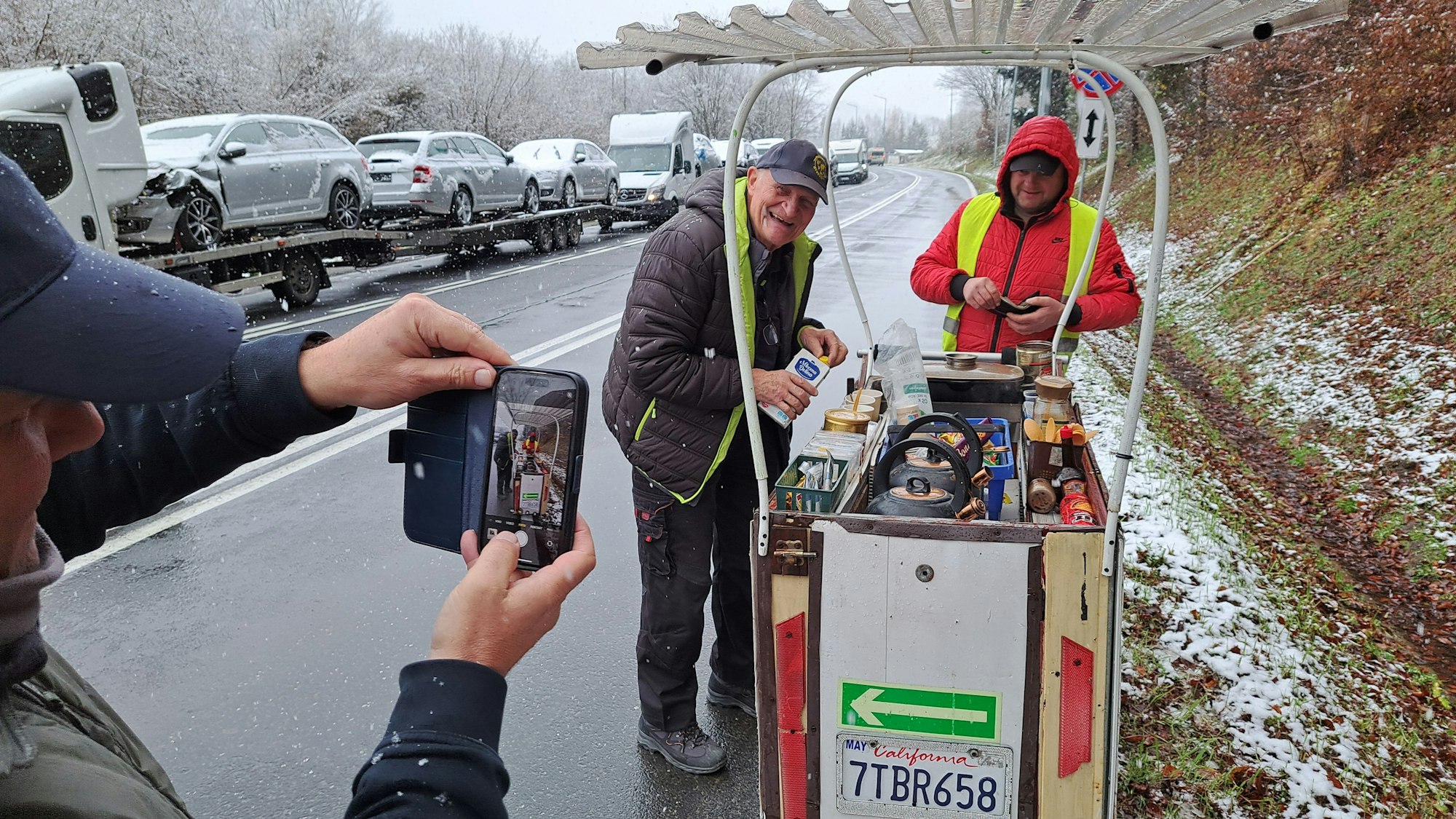 Ein Mann in Warnweste verkauft Tee und Kaffee von einem rollenden Verkaufswagen, links ist eine lange Autoschlange zu sehen.