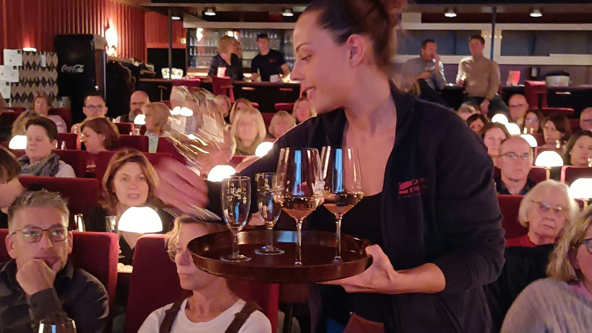 Zu sehen ist eine Servicekraft, die ein Tablett mit Weingläsern hält und im gut besuchten Kinosaal steht.