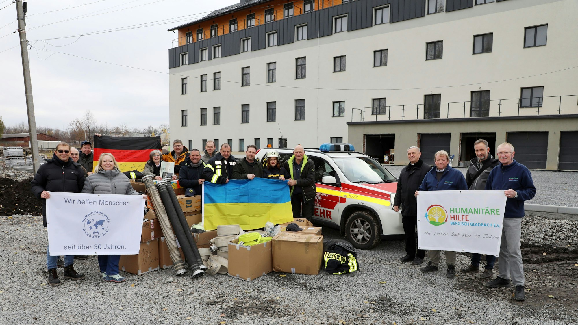 Menschen mit Transparenten und deutscher wie ukrainischer Fahne stehen zwischen Hilfsgütern und einem gespendeten Feuerwehrauto aus Kürten.