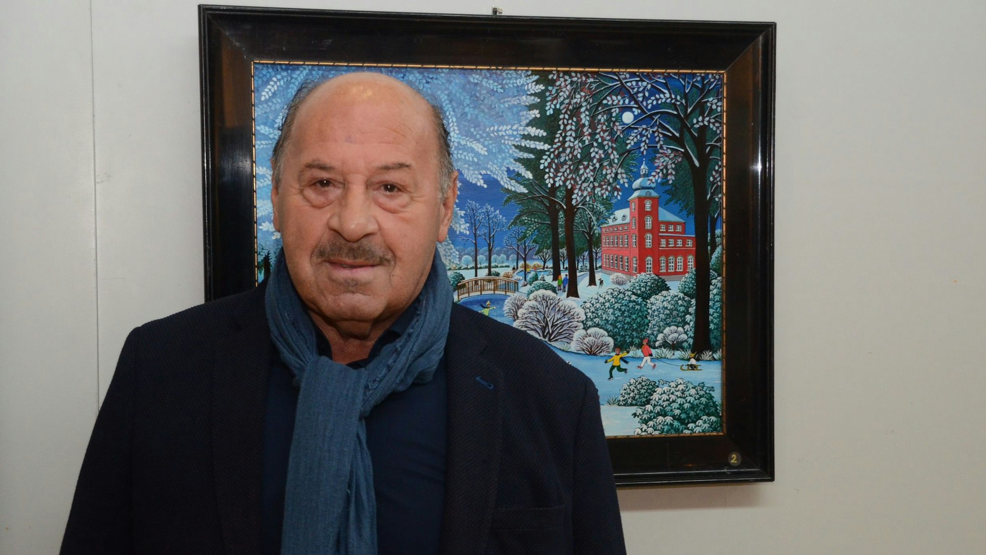 Ein Mann mit Schal und schwarzem Sakko steht vor einem naiven Gemälde.In einer Winterlandschaft leuchtet das rote Herrenhaus der Troisdorfer Burg Wissem..