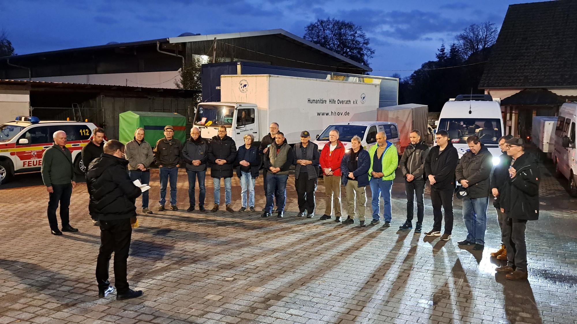 Fahrerinnen und Fahrer des 7. Hilfskonvois der Humanitären Hilfe Overath und Bergisch Gladbach stehen im Halbkreis vor den Konvoifahrzeugen, während Kaplan Michael Schiller ihnen den Reisesegen gibt.