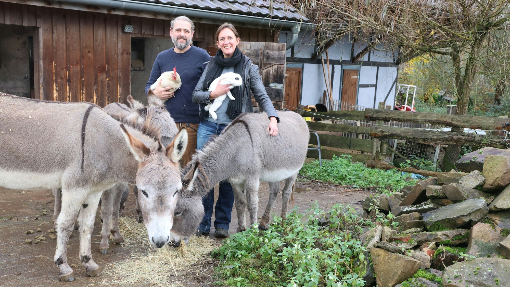 Ein Mann und eine Frau stehen auf einem Hof vor einer Scheune. Sie halten ein Kaninchen und ein Huhn in den Armen. Vor und neben ihnen stehen drei Esel.