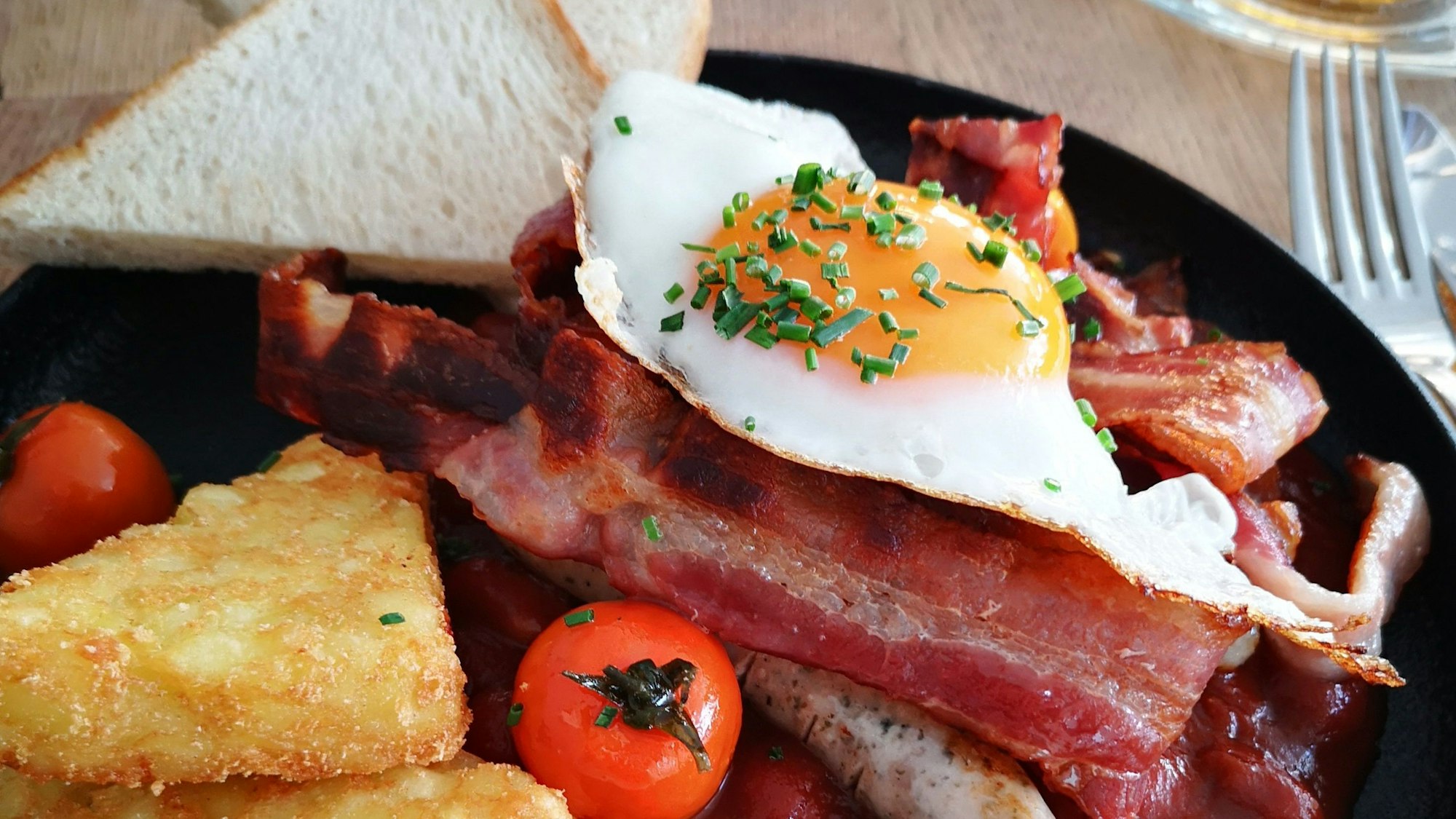 Auf einem Teller liegen Toastscheiben, ein Rösti, Cherrytomaten, gebratener Speck und ein Spiegelei.