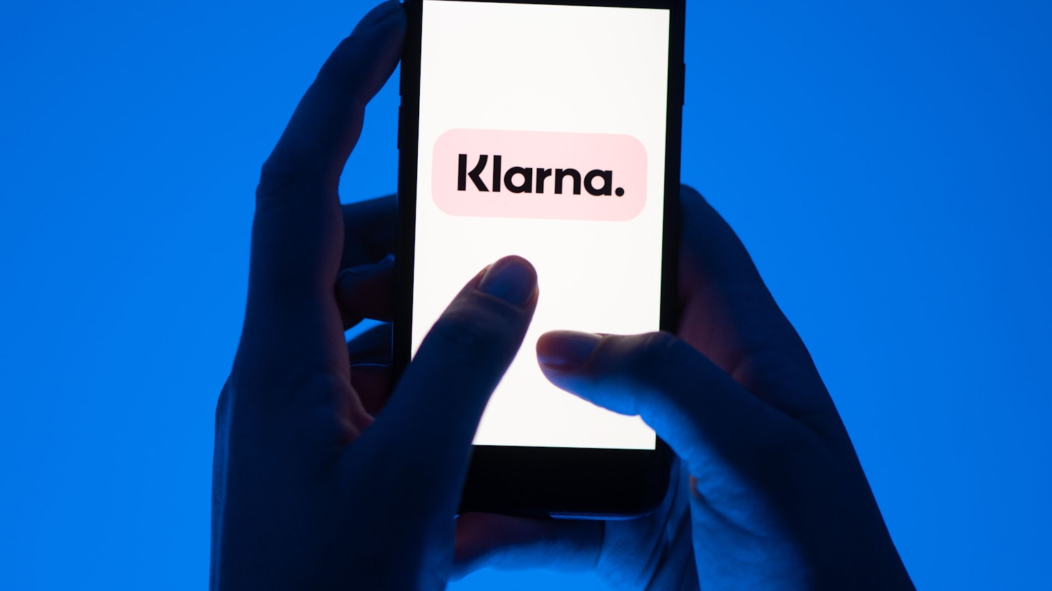 Zwei Hände halten ein Smartphone vor blauem Hintergrund, darauf ist groß das Klarna-Logo zu sehen.